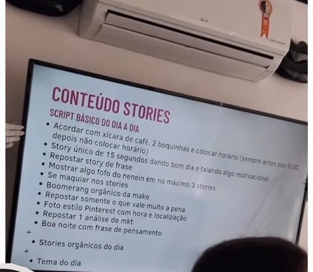 Imagem exemplificando o planejamento de uma agência de comunicação. Na imagem: um slide com tópicos é exibido numa tela de projeção, com um ar condicionado em cima.  No slide: "CONTEÚDO STORIES - SCRIPT BÁSICO DO DIA A DIA:
- Acordar com xícara de café, 2 boquinhas e colocar horário (sempre antes das 9:00, depois não colocar horário)
- Story único de 15 segundos dando bom dia e falando algo motivacional;
- Repostar story de frase;
' Mostrar algo fofo do nenein em no máximo 3 stories
- Se maquiar nos stories
- Boomerang orgânico da make
- Respostar somente o que vale muito a pena
- Foto estilo Pinterest com hora e localização
- Repostar 1 análise de mkt
- Boa noite com frase de pensamento
+
- Stories orgânicos do dia
+
- Tema do dia
